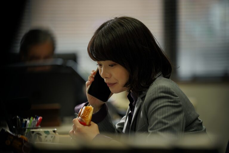 映画『佐藤さんと佐藤さん』よりデスクでパンをほおばるサチ（岸井ゆきの）