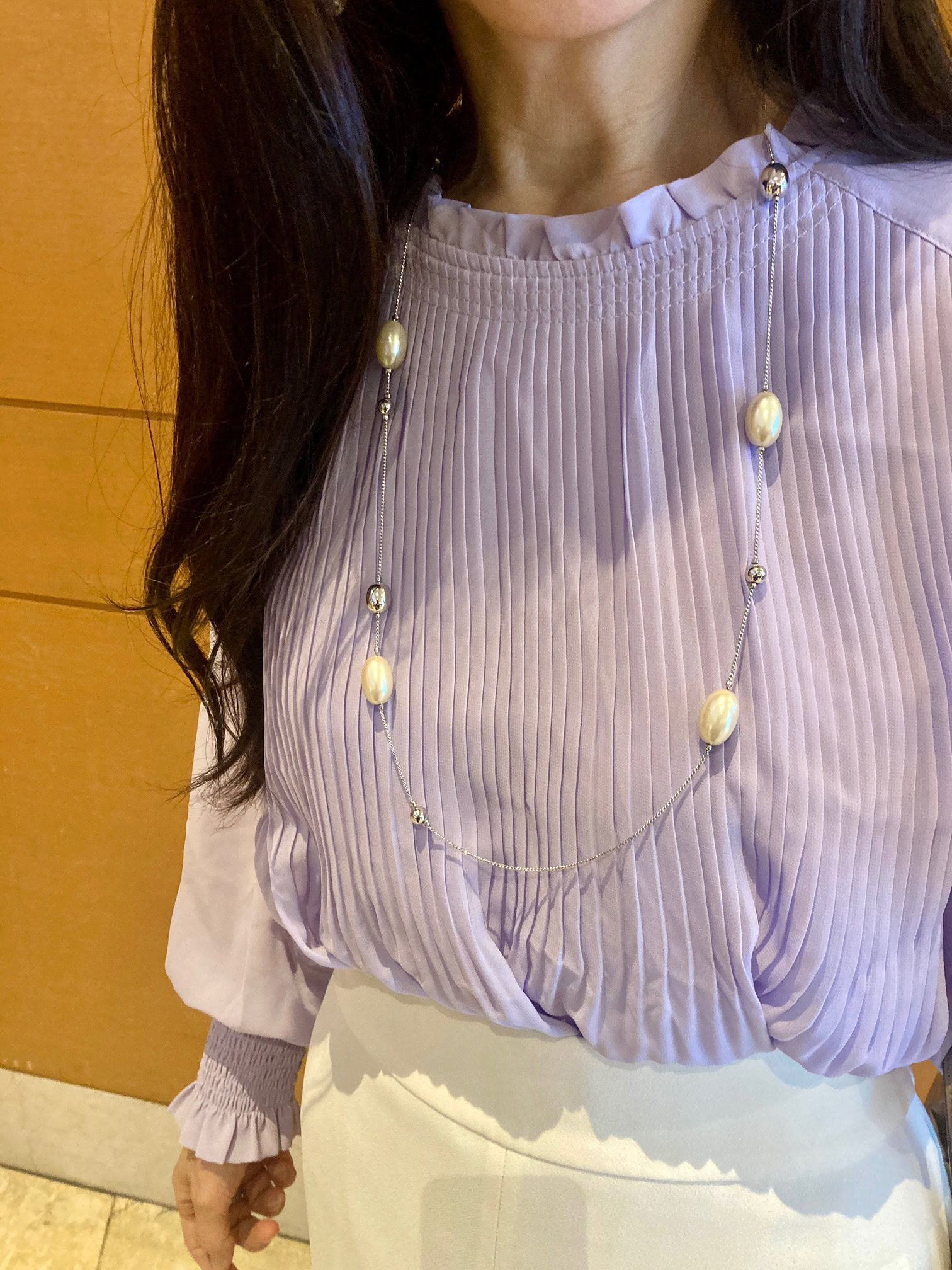 「VIS」のシアーなプリーツ素材に首元＆袖口のフリルが愛らしい、淡いラベンダーのブラウス。「首元のフリルと、パールとシルバーのロングネックレスで、華やかさを添えました」